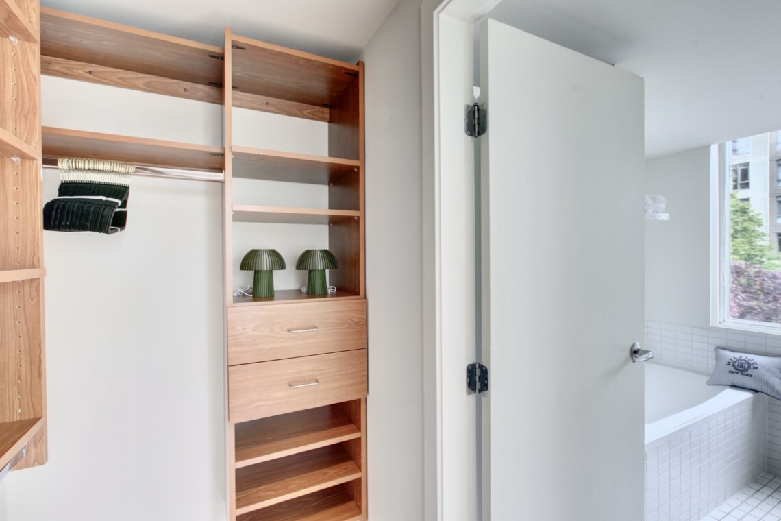 Open closet with tall wooden shelves and drawers, two green ribbed lamps on a shelf, and a doorway to a bright bathroom.