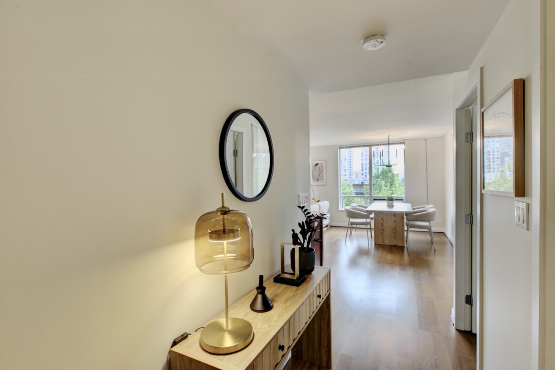 View down a bright hallway into a living/dining area with a wooden console table, gold lamp, round mirror, and potted plants against light walls and hardwood floors.