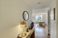View down a bright hallway into a living/dining area with a wooden console table, gold lamp, round mirror, and potted plants against light walls and hardwood floors.