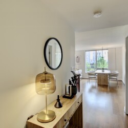 View down a bright hallway into a living/dining area with a wooden console table, gold lamp, round mirror, and potted plants against light walls and hardwood floors.