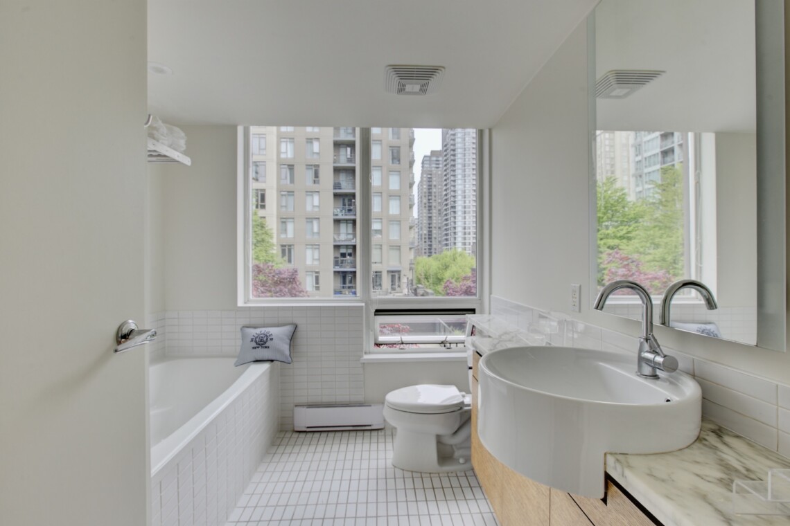 Bright modern bathroom with a white tub, toilet, and curved sink, large window showing city buildings outside