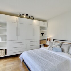 Modern white bedroom with a full wall of built-in wardrobes, open shelves, and a bed with white bedding on a wooden floor.