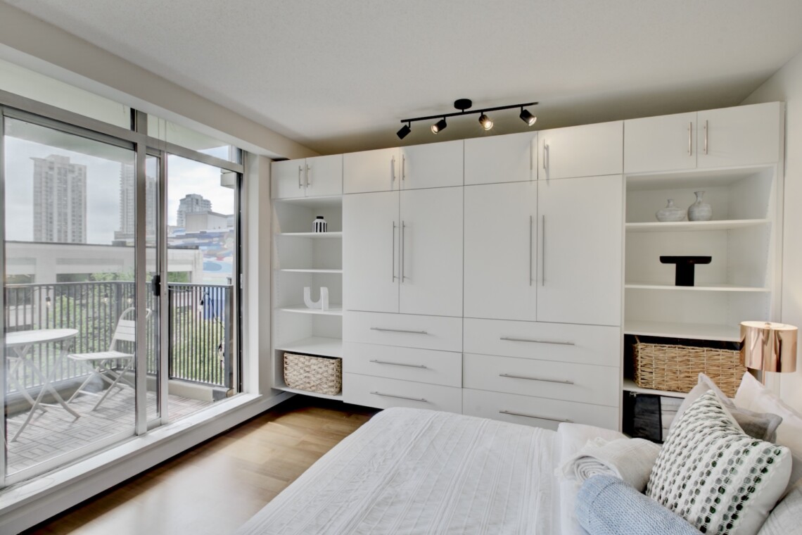 Modern white built-in wardrobe with open shelves and baskets, beside glass balcony doors showing a cityscape nearby.