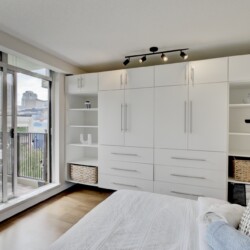Modern white built-in wardrobe with open shelves and baskets, beside glass balcony doors showing a cityscape nearby.