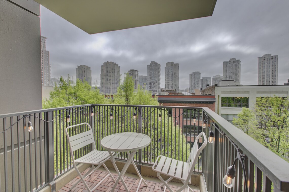 Balcony with white metal table and chairs, string lights along the railing, overlooking green trees and tall gray city buildings under a cloudy sky.