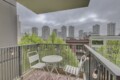 Balcony with white metal table and chairs, string lights along the railing, overlooking green trees and tall gray city buildings under a cloudy sky.
