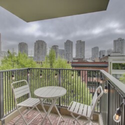 Balcony with white metal table and chairs, string lights along the railing, overlooking green trees and tall gray city buildings under a cloudy sky.