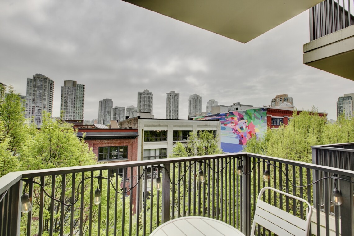 Balcony railing with string lights overlooks a cityscape of tall buildings and a colorful mural on a brick building, under overcast skies.