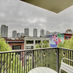 Balcony railing with string lights overlooks a cityscape of tall buildings and a colorful mural on a brick building, under overcast skies.