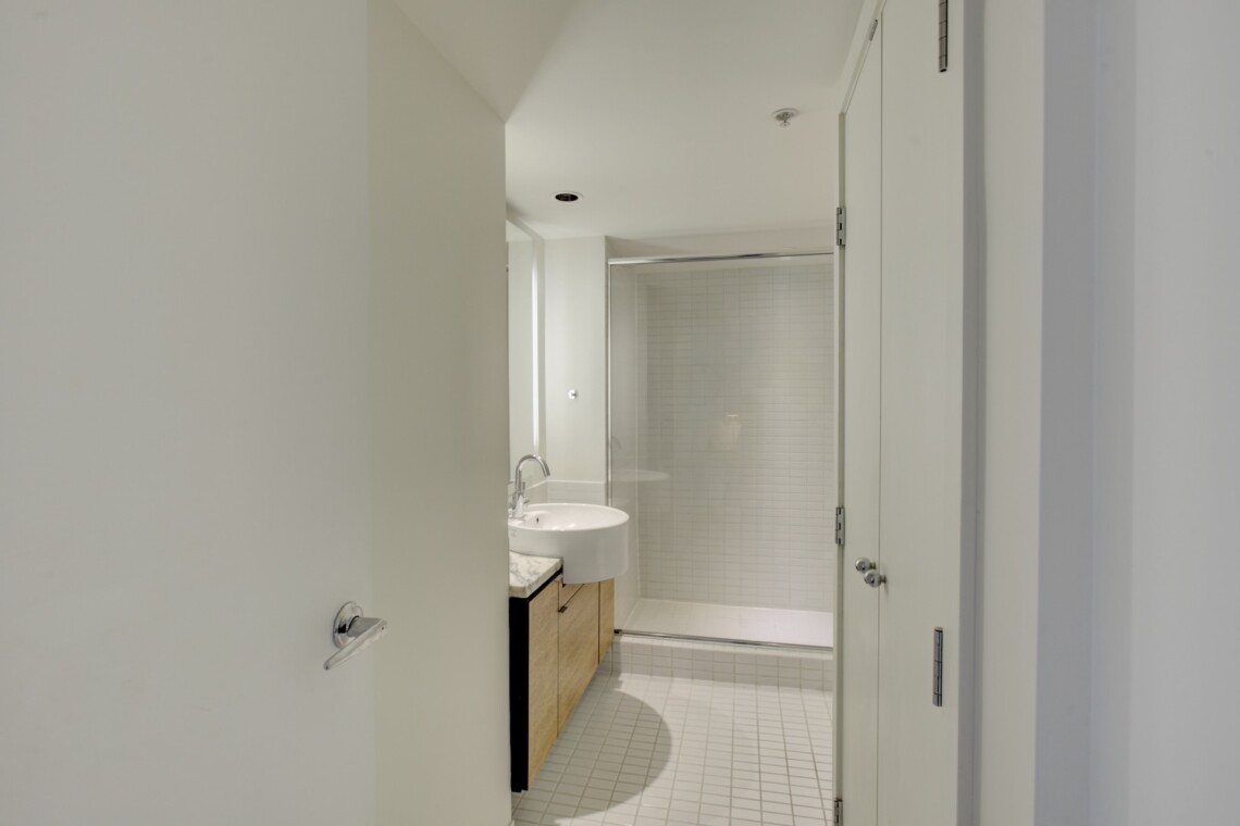 Modern bathroom interior with a glass shower enclosure, white tiles, and a light wood vanity under bright overhead lighting