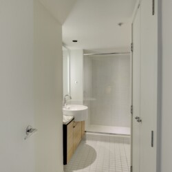 Modern bathroom interior with a glass shower enclosure, white tiles, and a light wood vanity under bright overhead lighting