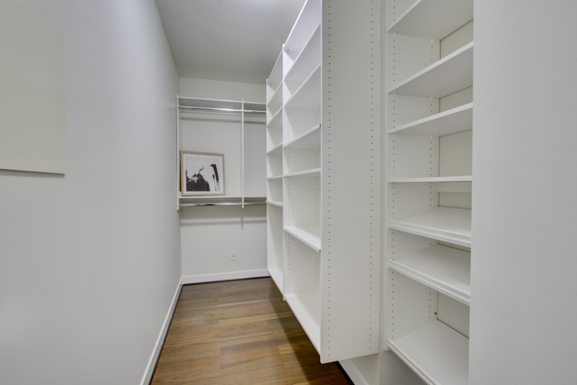 Narrow walk-in closet with white open shelves, a metal clothing rod, and a framed artwork on the back wall.