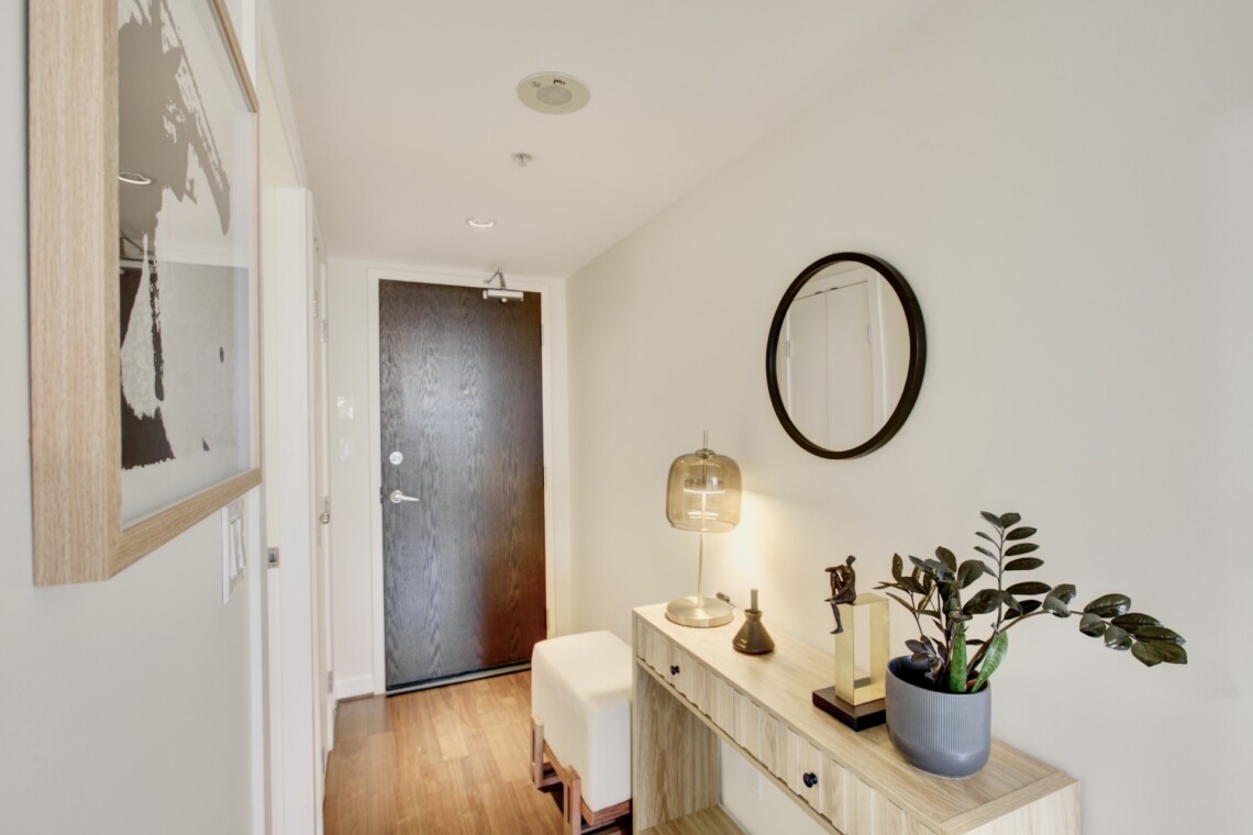 Entryway with a dark front door, light wood console table, round black-framed mirror, and a table lamp beside a plant.