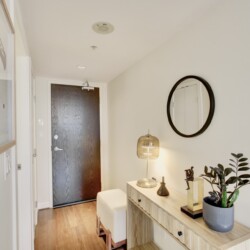 Entryway with a dark front door, light wood console table, round black-framed mirror, and a table lamp beside a plant.