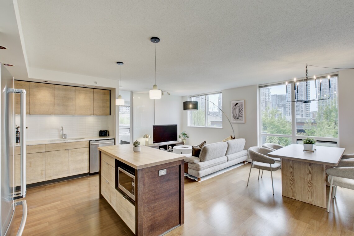 Open-concept living area with a kitchen on the left, a wooden island, a sofa, and large windows overlooking a cityscape outside.