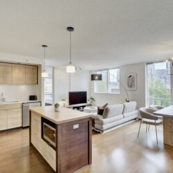 Open-concept living area with a kitchen on the left, a wooden island, a sofa, and large windows overlooking a cityscape outside.