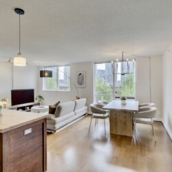 Open-concept living space with a kitchen island, dining table, and beige sofas, illuminated by large windows and pendant lights.