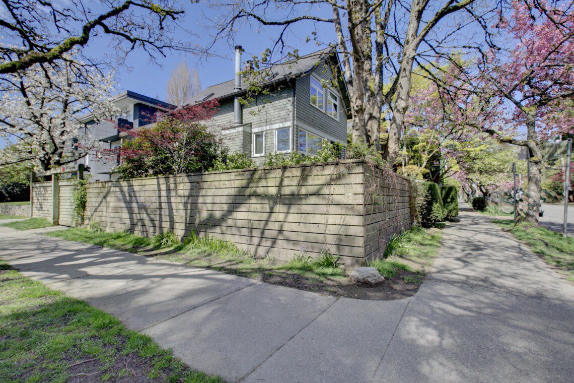 Two-story house behind a tall wooden fence, with blossoming trees along the sidewalk under a clear blue sky.