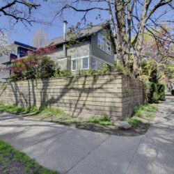 Two-story house behind a tall wooden fence, with blossoming trees along the sidewalk under a clear blue sky.