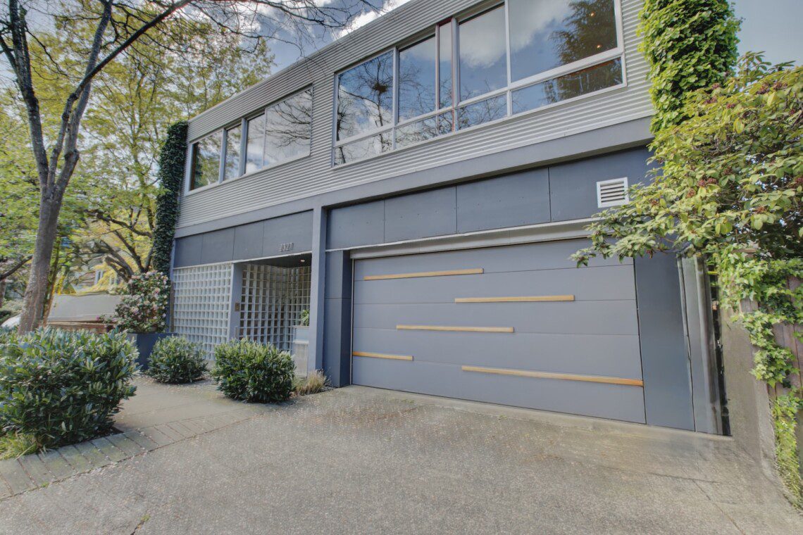 Modern two-story blue-gray building with large glass windows and a wide garage door featuring horizontal wooden slats, surrounded by greenery.