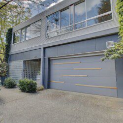 Modern two-story blue-gray building with large glass windows and a wide garage door featuring horizontal wooden slats, surrounded by greenery.