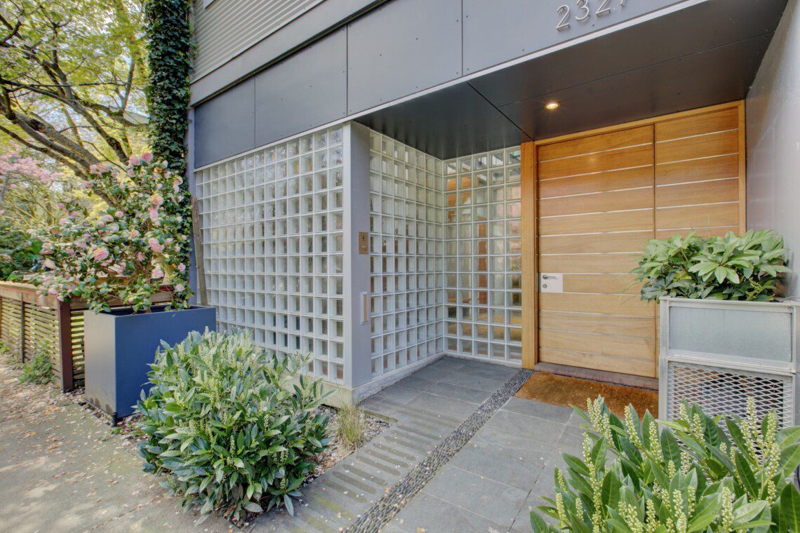 Modern building entrance with a glass block wall, wood-paneled door, and lush green plants along the walkway.