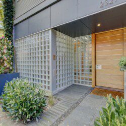 Modern building entrance with a glass block wall, wood-paneled door, and lush green plants along the walkway.