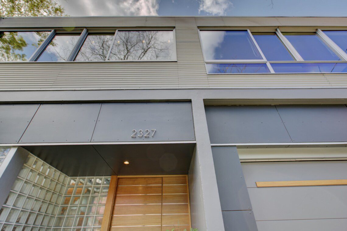 Modern building entrance with grey metal panels, large windows reflecting the sky, and a wooden door under a recessed canopy; address 2327.