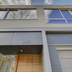 Modern building entrance with grey metal panels, large windows reflecting the sky, and a wooden door under a recessed canopy; address 2327.