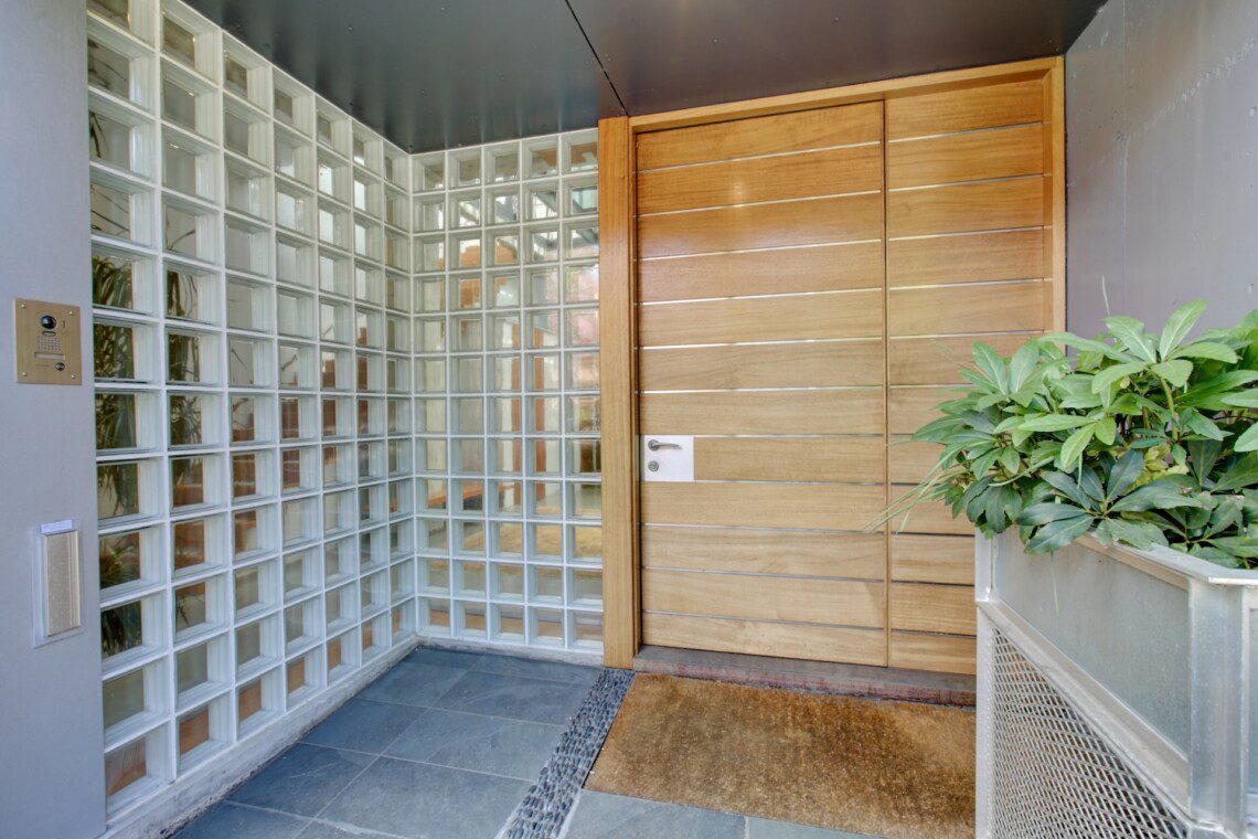 Modern entryway with a tall wooden paneled door beside a glass block wall and potted greenery on the right