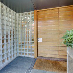 Modern entryway with a tall wooden paneled door beside a glass block wall and potted greenery on the right