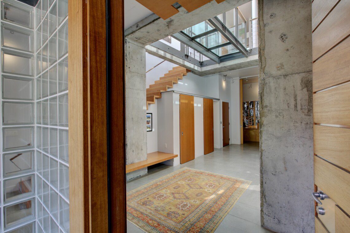 Open-concept foyer with concrete columns, a floating wooden staircase, and multiple wooden doors along a white wall.