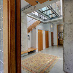 Open-concept foyer with concrete columns, a floating wooden staircase, and multiple wooden doors along a white wall.