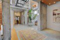 Open industrial lobby with concrete columns, a large leafy plant in a white pot, and a colorful rug near a wooden bench; office area visible in back.