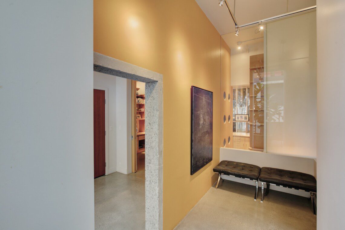 Modern interior hallway with an orange accent wall, a large chalkboard, and black tufted benches along a glass partition.