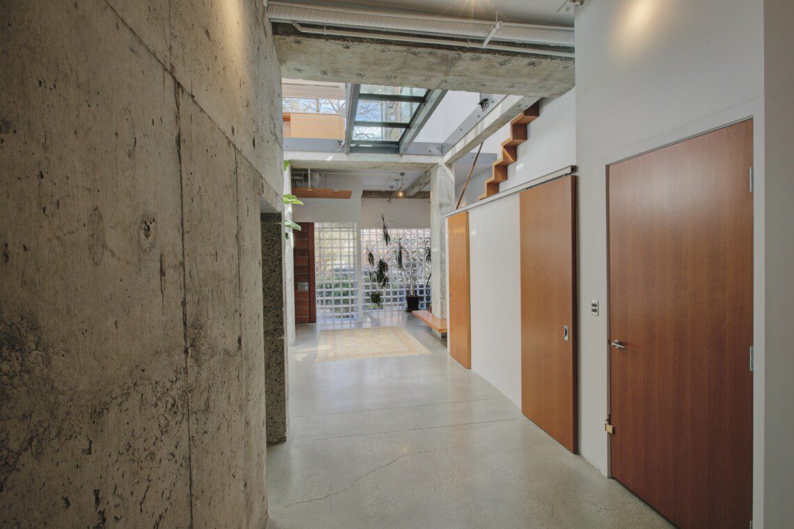 Industrial-style hallway with concrete walls, wooden doors on the right, and a glass ceiling letting in natural light.