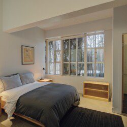 Bedroom with a queen bed, gray bedding, two wooden nightstands, and a large window showing leafless trees outside.