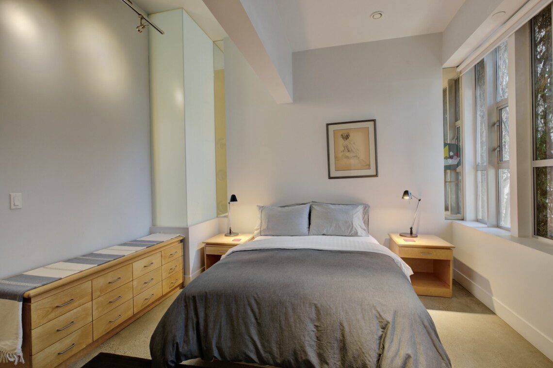 Bedroom with a centered bed, gray bedding, two wooden nightstands with lamps, and a framed drawing above the headboard.