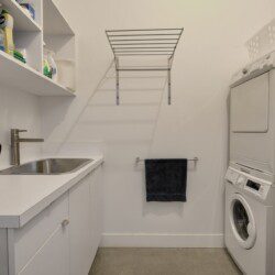 Laundry room with a sink, counters, and white cabinets, plus a stacked washer/dryer on the right and a shelf of cleaners above.