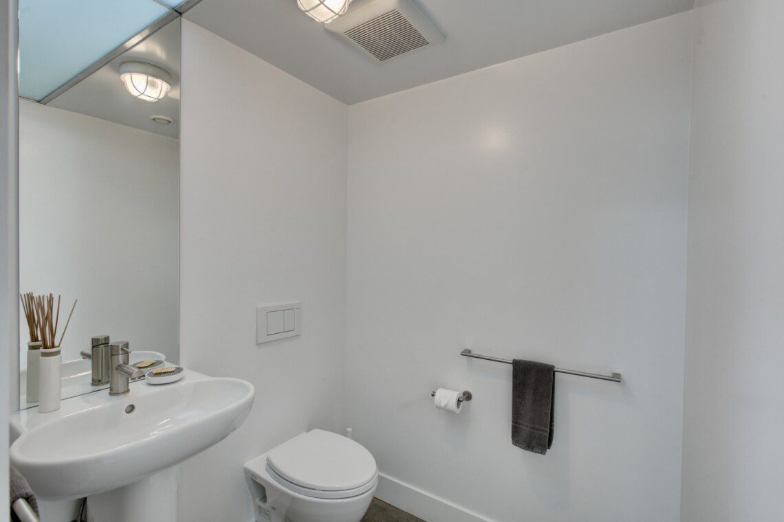 Small white bathroom with pedestal sink, mirror, and wall-mounted toilet; towel bar with a dark towel on the right.