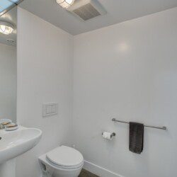 Small white bathroom with pedestal sink, mirror, and wall-mounted toilet; towel bar with a dark towel on the right.