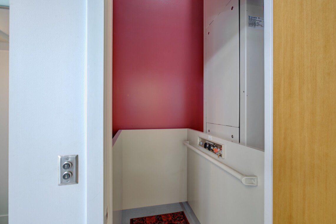 Interior of a small elevator cab with a red back wall, white side panels, and a control panel with colorful buttons on the right; a handrail along the lower wall.