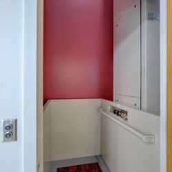 Interior of a small elevator cab with a red back wall, white side panels, and a control panel with colorful buttons on the right; a handrail along the lower wall.