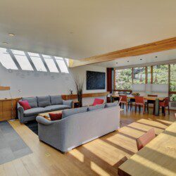 Bright open-plan living and dining area with gray couches, wooden beams, and large windows overlooking greenery; natural light fills the space.
