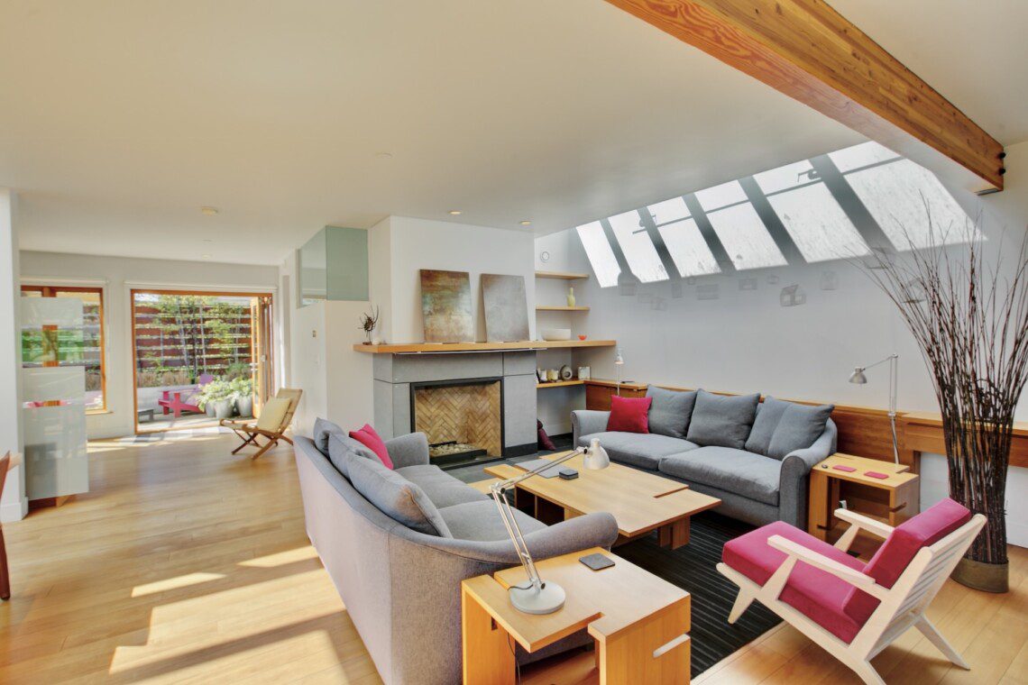 Bright, modern living room with a skylight and wooden beam, gray sofas, and pink accents around a central fireplace.