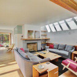Bright, modern living room with a skylight and wooden beam, gray sofas, and pink accents around a central fireplace.