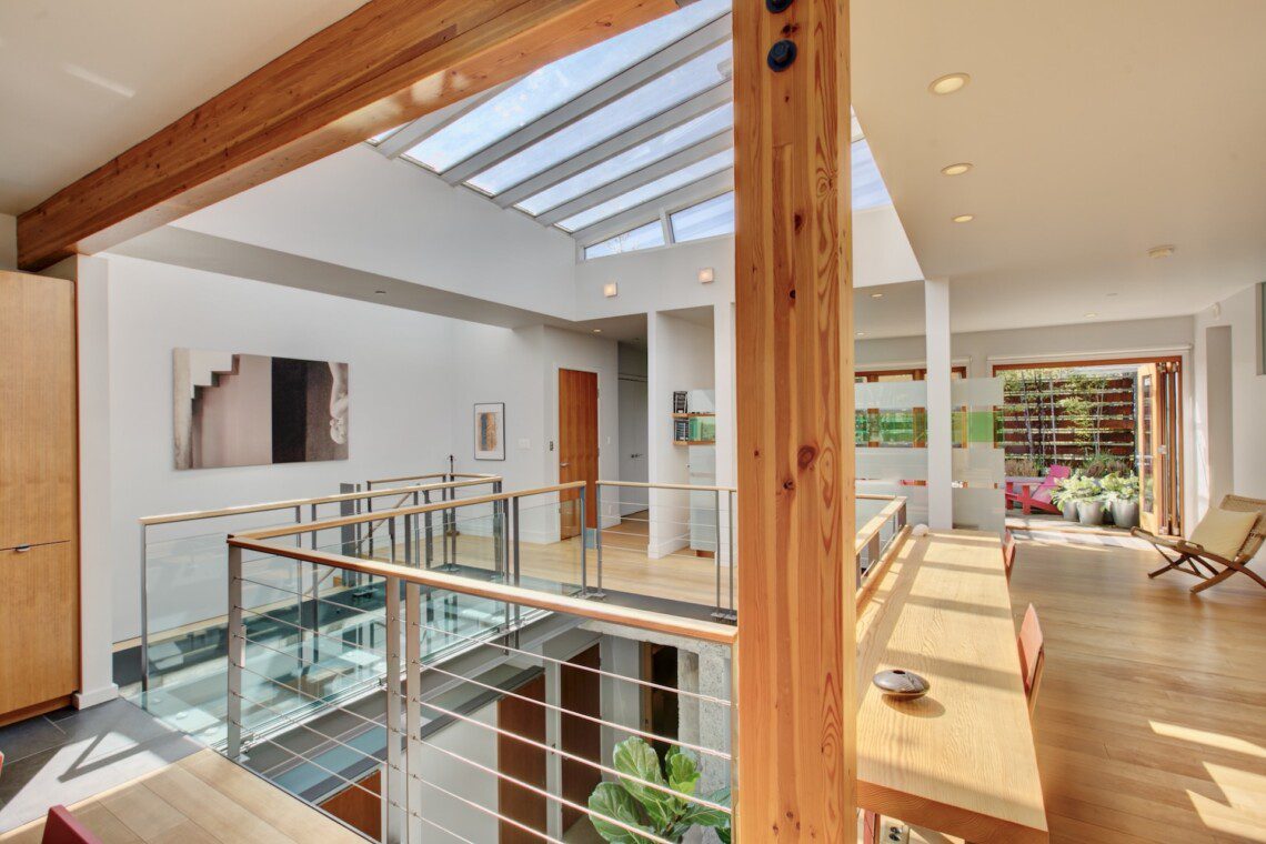 Bright open living area with a skylight, wooden beams, and glass railing along a mezzanine walkway.