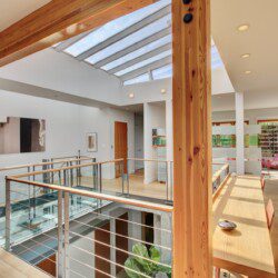 Bright open living area with a skylight, wooden beams, and glass railing along a mezzanine walkway.
