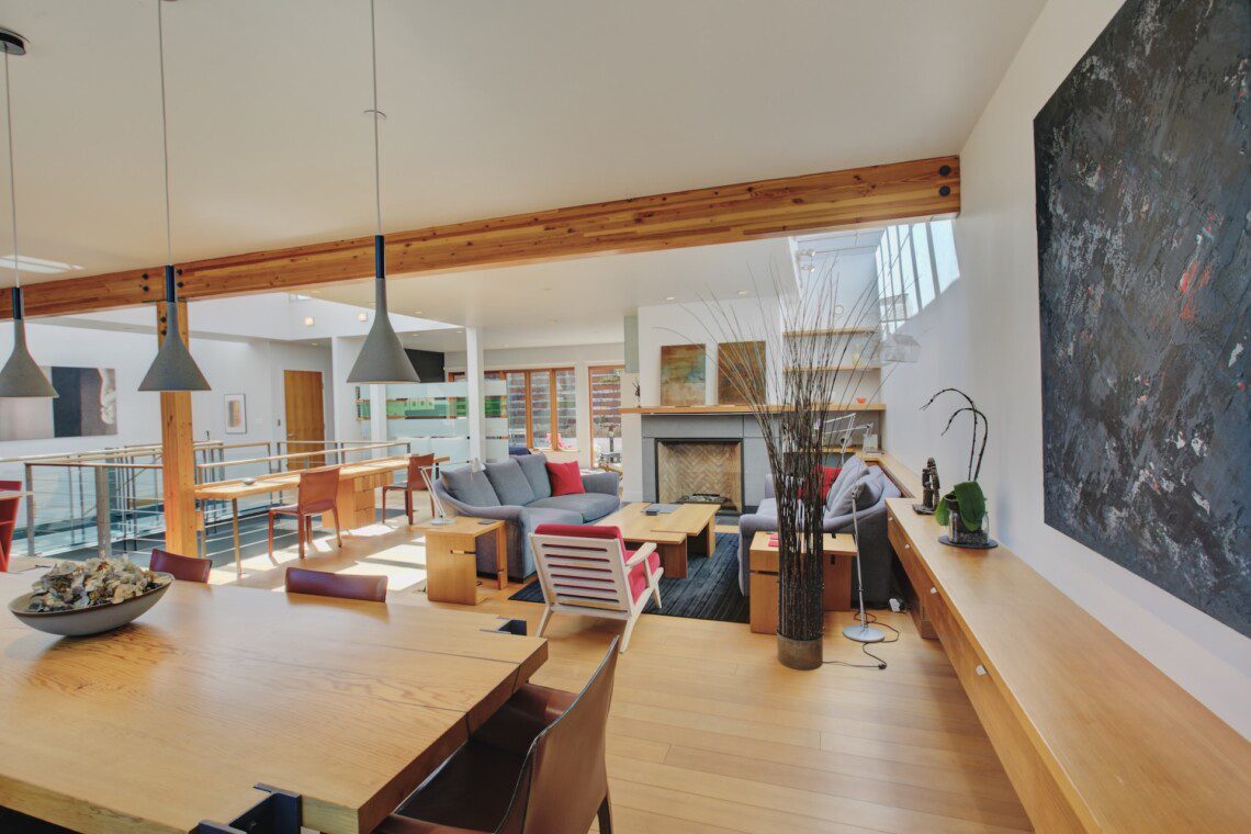 Open living space with wooden floors, ceiling beams, a gray sectional sofa, and a fireplace in the center