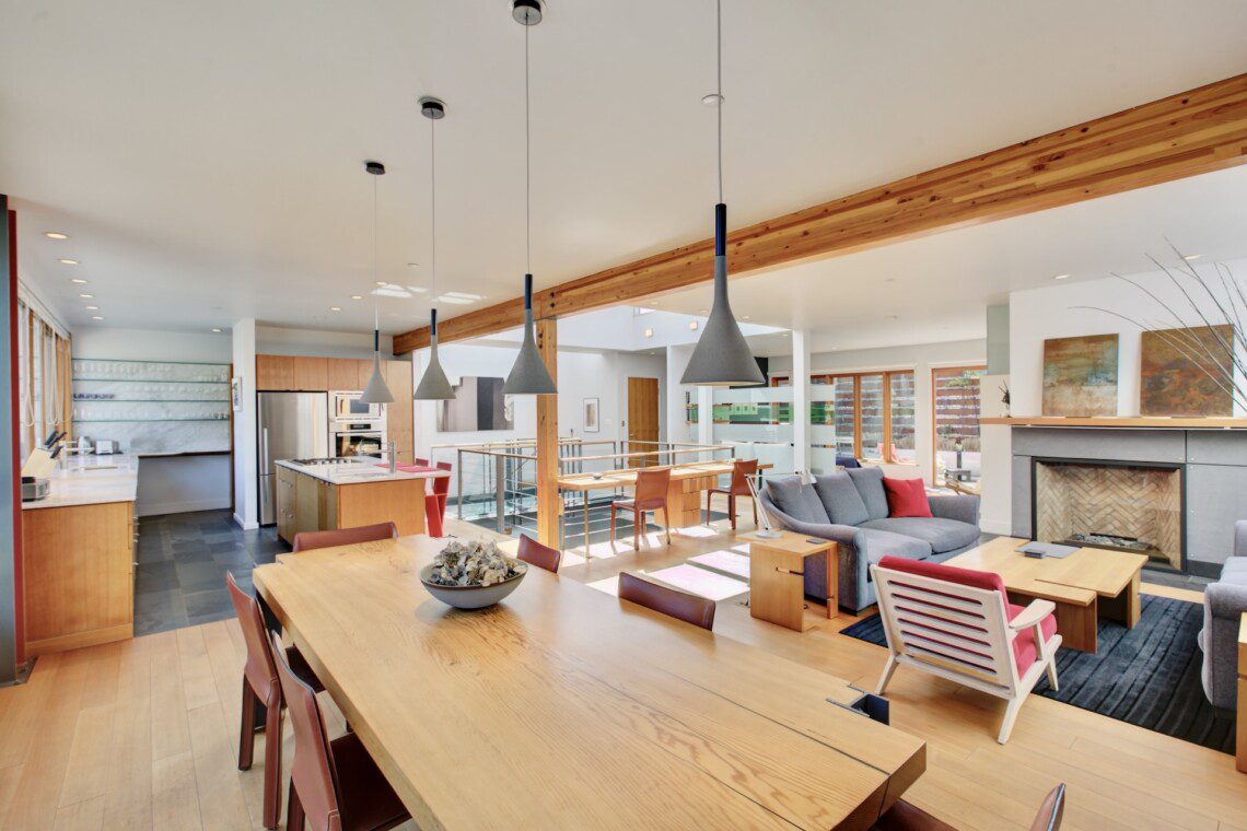 Open-concept living space with a long wooden dining table, pendant lights, and a kitchen area with stainless appliances alongside a gray sofa and fireplace.